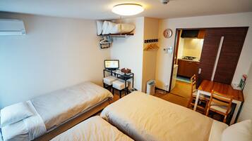 A compact hotel room with two single beds, a small desk with a computer, a chair, and a kitchenette area.