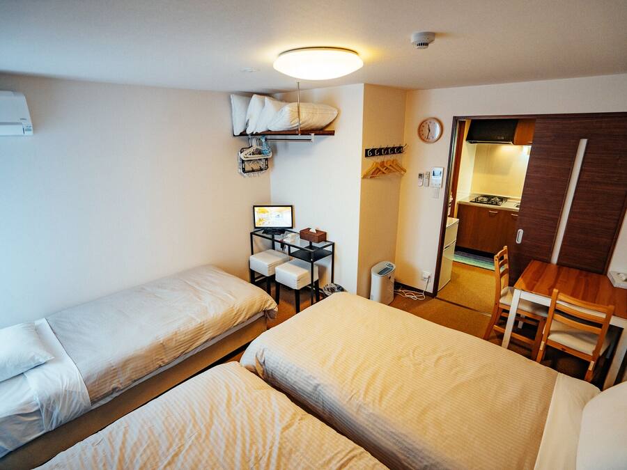 A compact hotel room with two single beds, a small desk with a computer, a chair, and a kitchenette area.