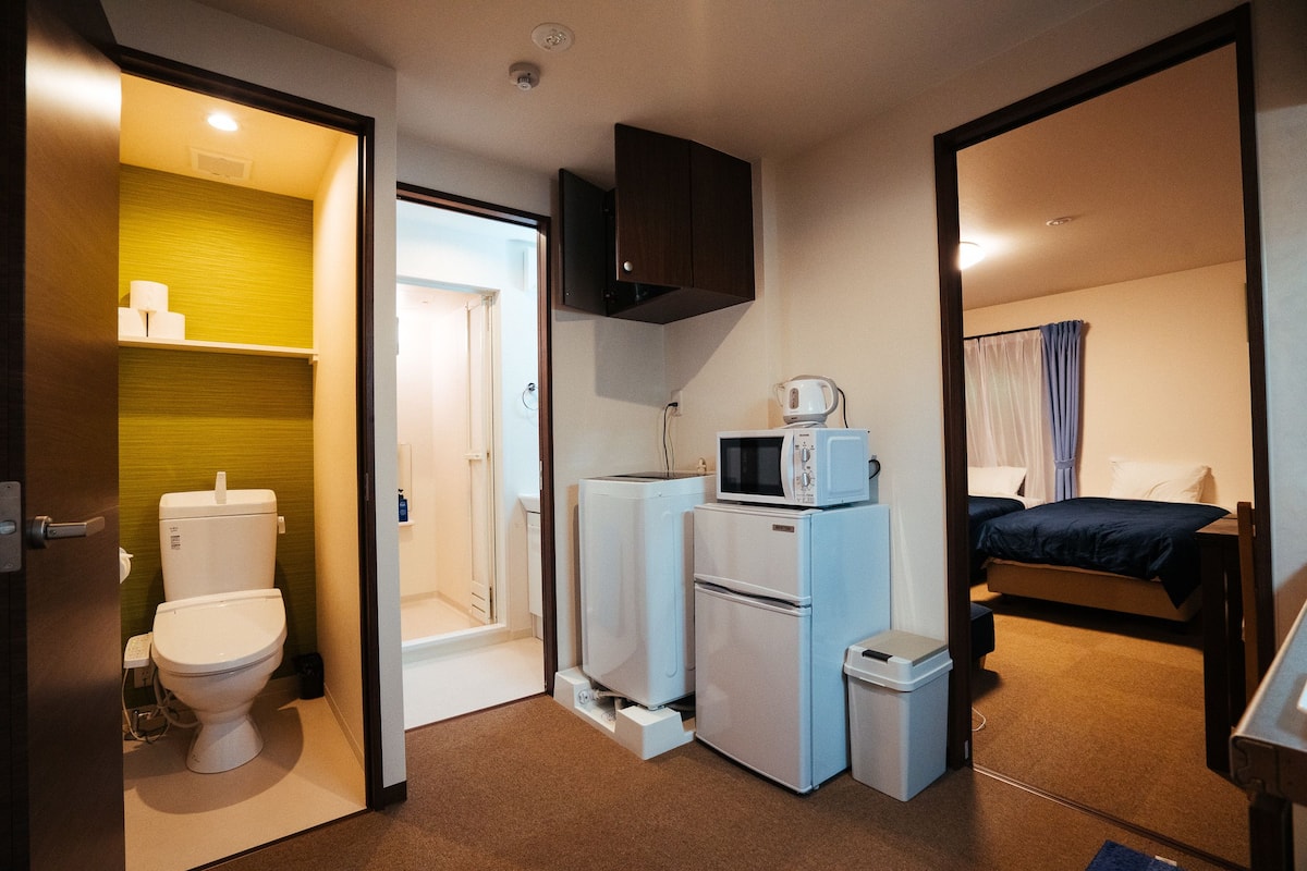 A compact hotel room with a toilet, a small kitchenette with a microwave, a refrigerator, and a bed with a blue blanket.