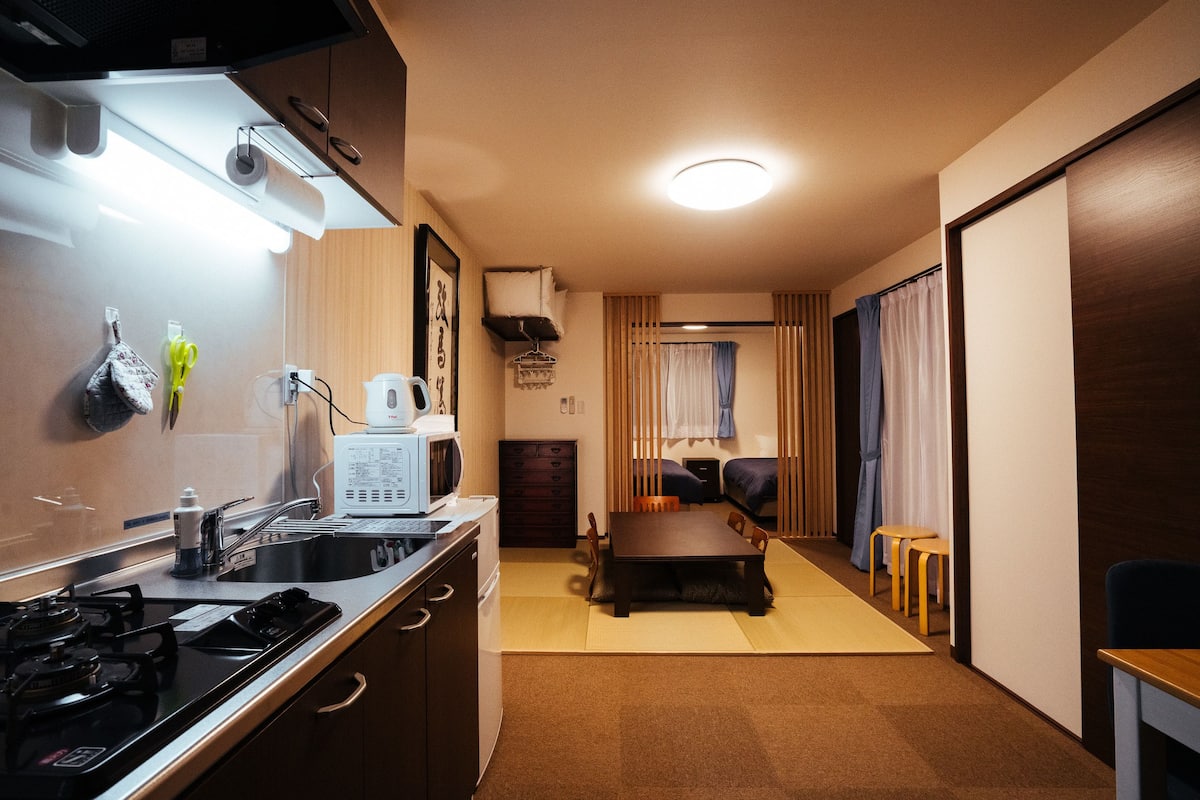 A compact kitchen with a stove, microwave, and sink. There is a dining area with a table and chairs, and a small seating area with a sofa and curtains.