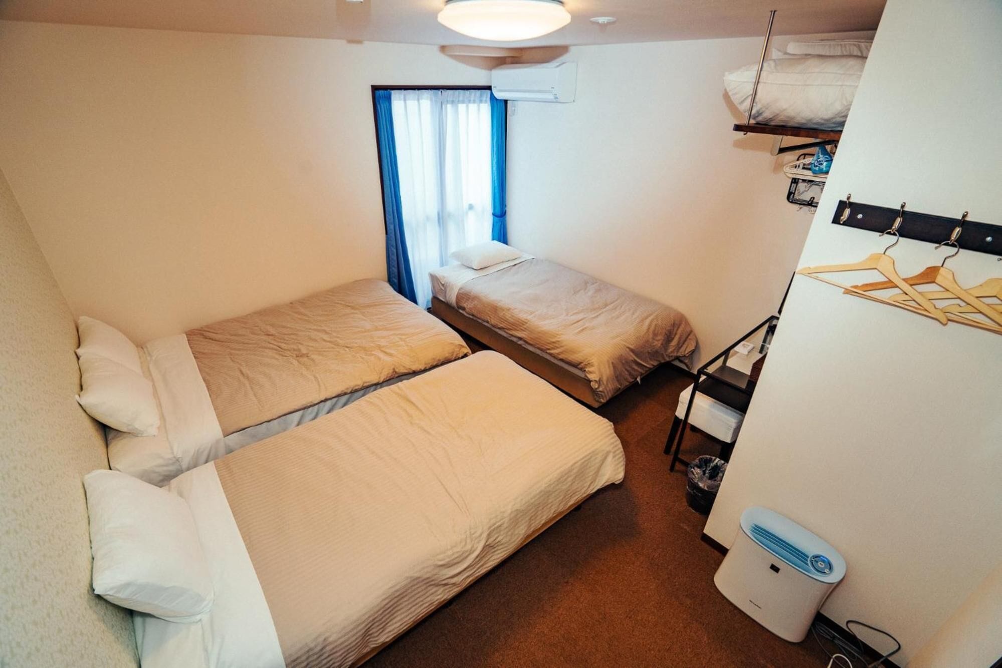 Une chambre de taille modeste, équipée de deux lits simples, d’une fenêtre avec des rideaux bleus, d’un ventilateur de plafond et d’un petit radiateur.