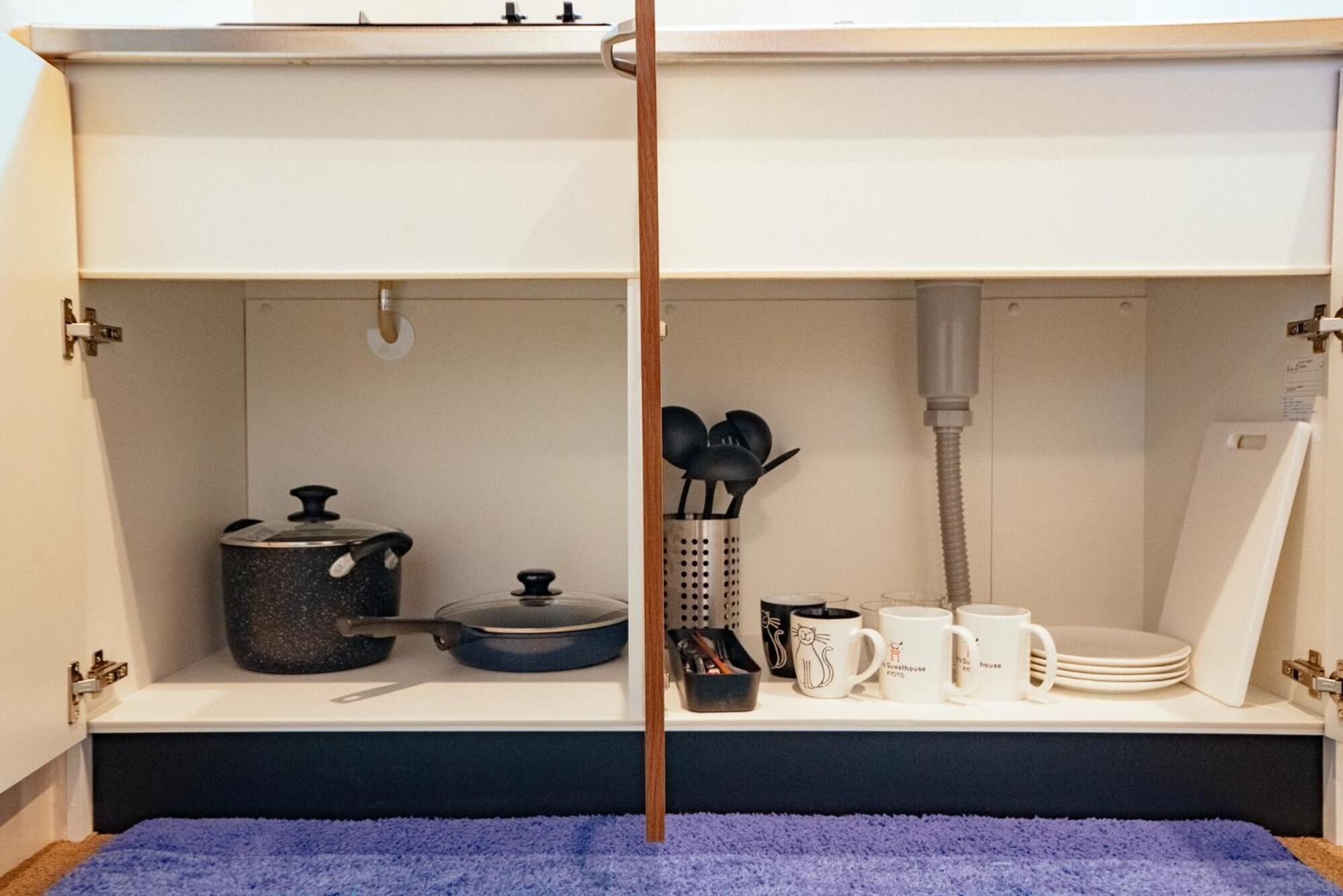 A kitchen with a blue towel on the floor, a pot, a colander, a coffee maker, and a stack of plates.