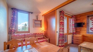A cozy cabin interior with a wooden dining table, benches, a small kitchen area, and a window with curtains. - Cozy studio 80m from lifts and 100m from shops in Valmorel (Valmorel)