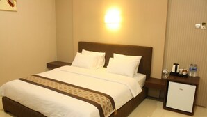 Cama bem arrumada com lençóis brancos e cabeceira marrom, mesa de cabeceira com abajur e um pequeno frigorífico.