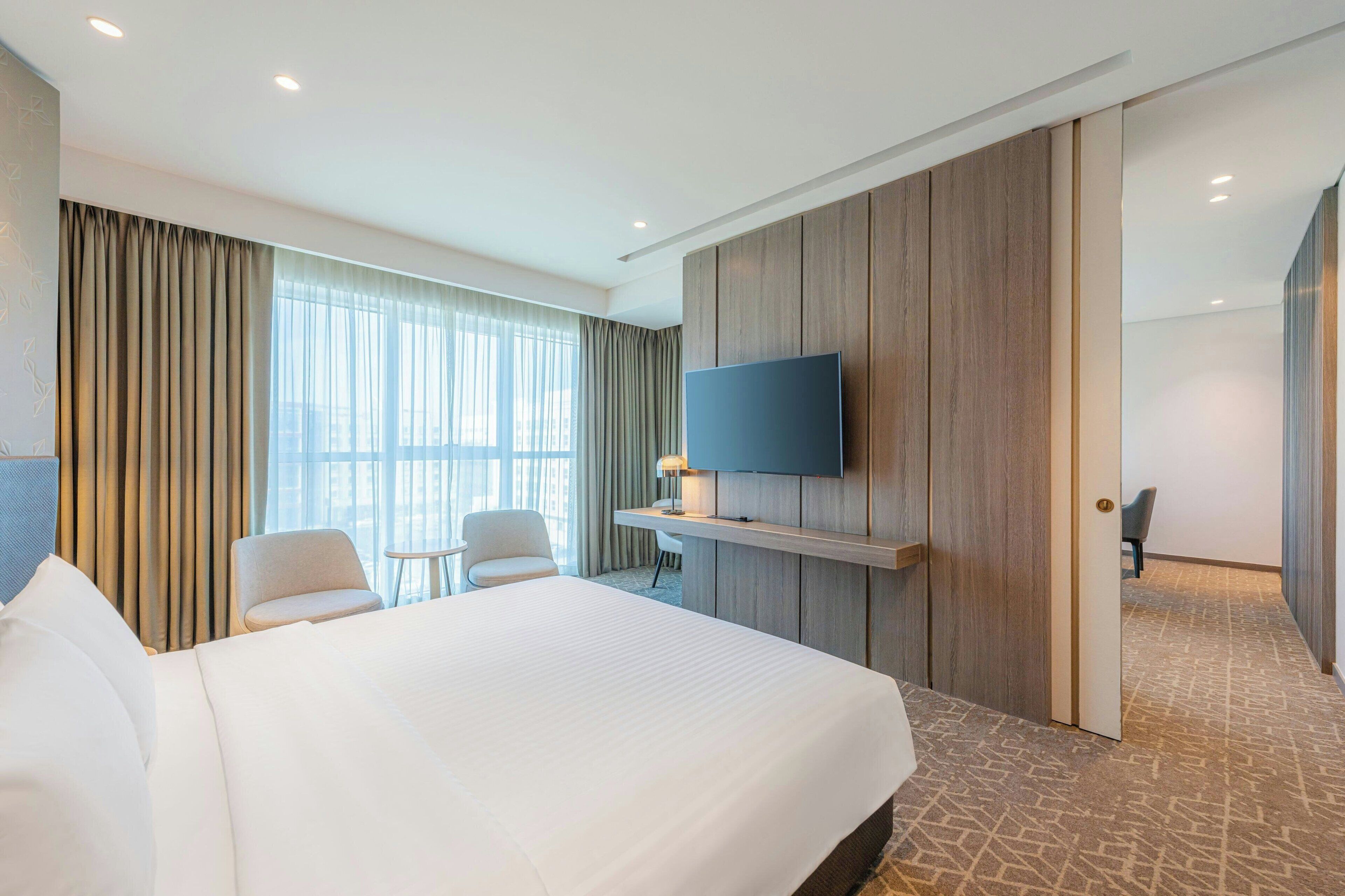 A hotel room with a large bed, a desk with a chair, a TV mounted on the wall, and a view of the city through large windows.