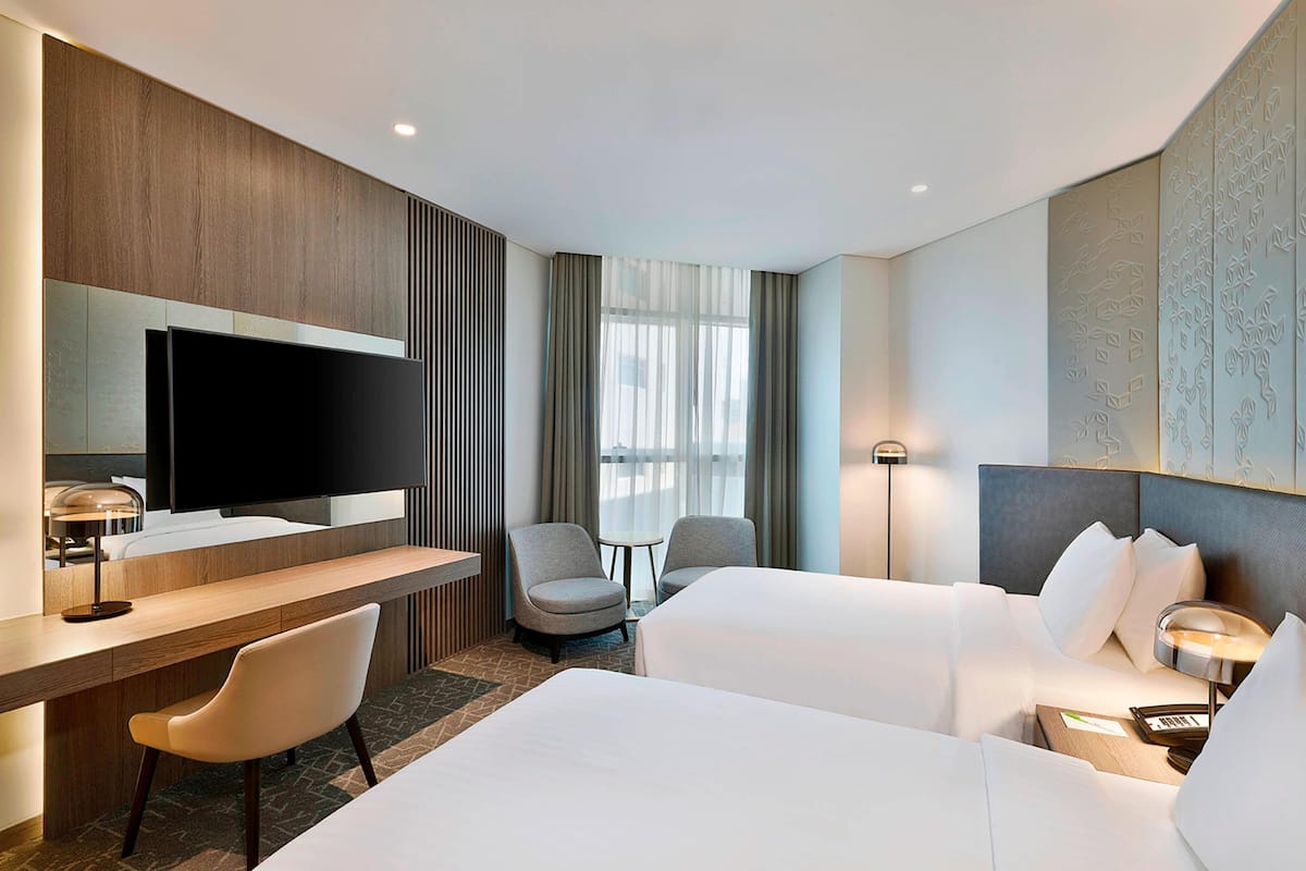 A hotel room with a flat-screen TV, a desk with a lamp, two chairs, a bed with white linens, and a small table with a lamp.