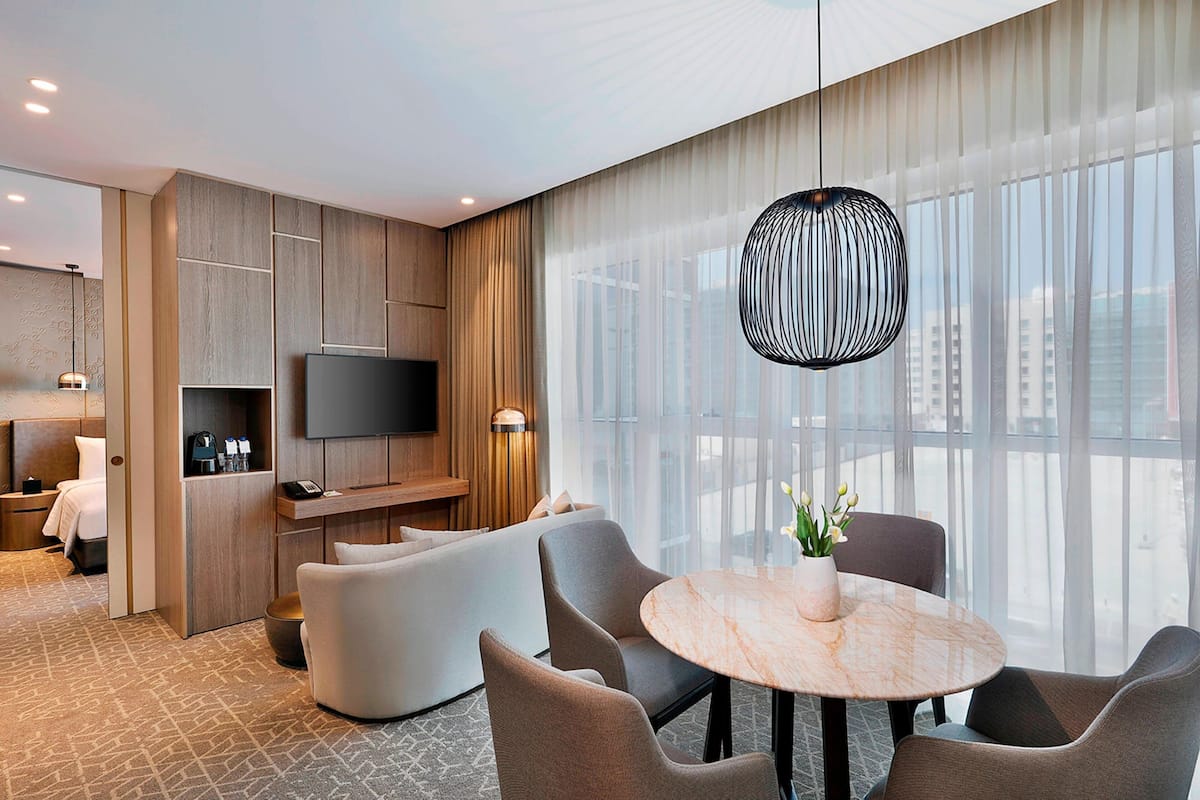 A modern hotel room with a sofa, a dining table, a TV, and a large window with curtains.