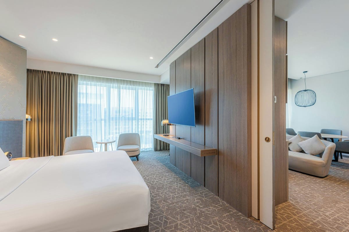 A hotel room with a large bed, a TV mounted on a wooden panel, a seating area with a sofa and chairs, and a view of the city through large windows.
