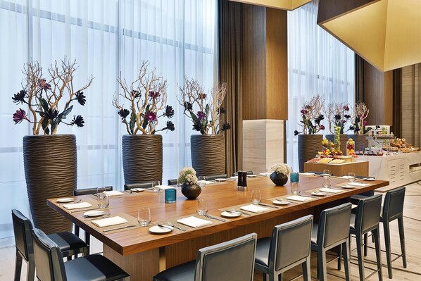 A conference room with a long wooden table set for a meeting, surrounded by chairs. There are large potted plants with flowers and branches, and a buffet area with various food items. - Courtyard by Marriott Al Barsha, Dubai (Dubai)