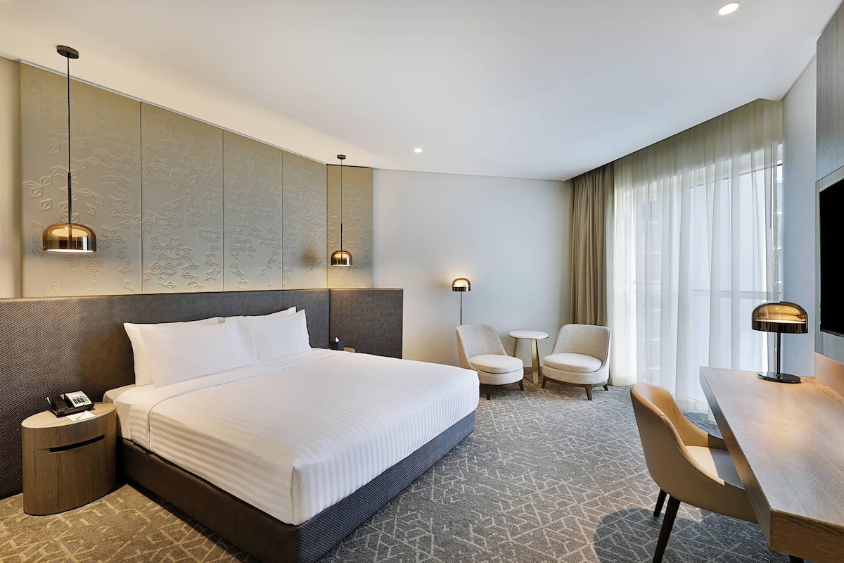 A hotel room with a large bed, bedside tables, a desk, chairs, a TV, and a window with curtains.