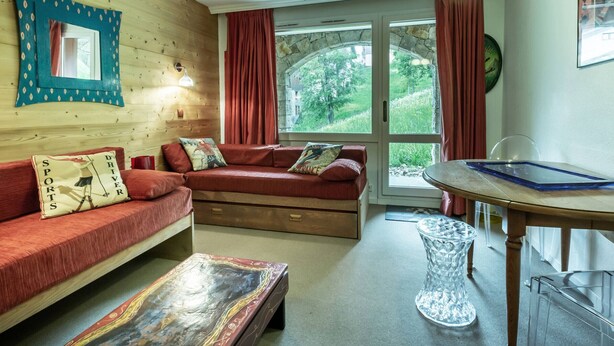 Ein gemütliches Wohnzimmer mit einer Holzwand, einem roten Sofa, einem Glastisch und Blick ins Grüne.