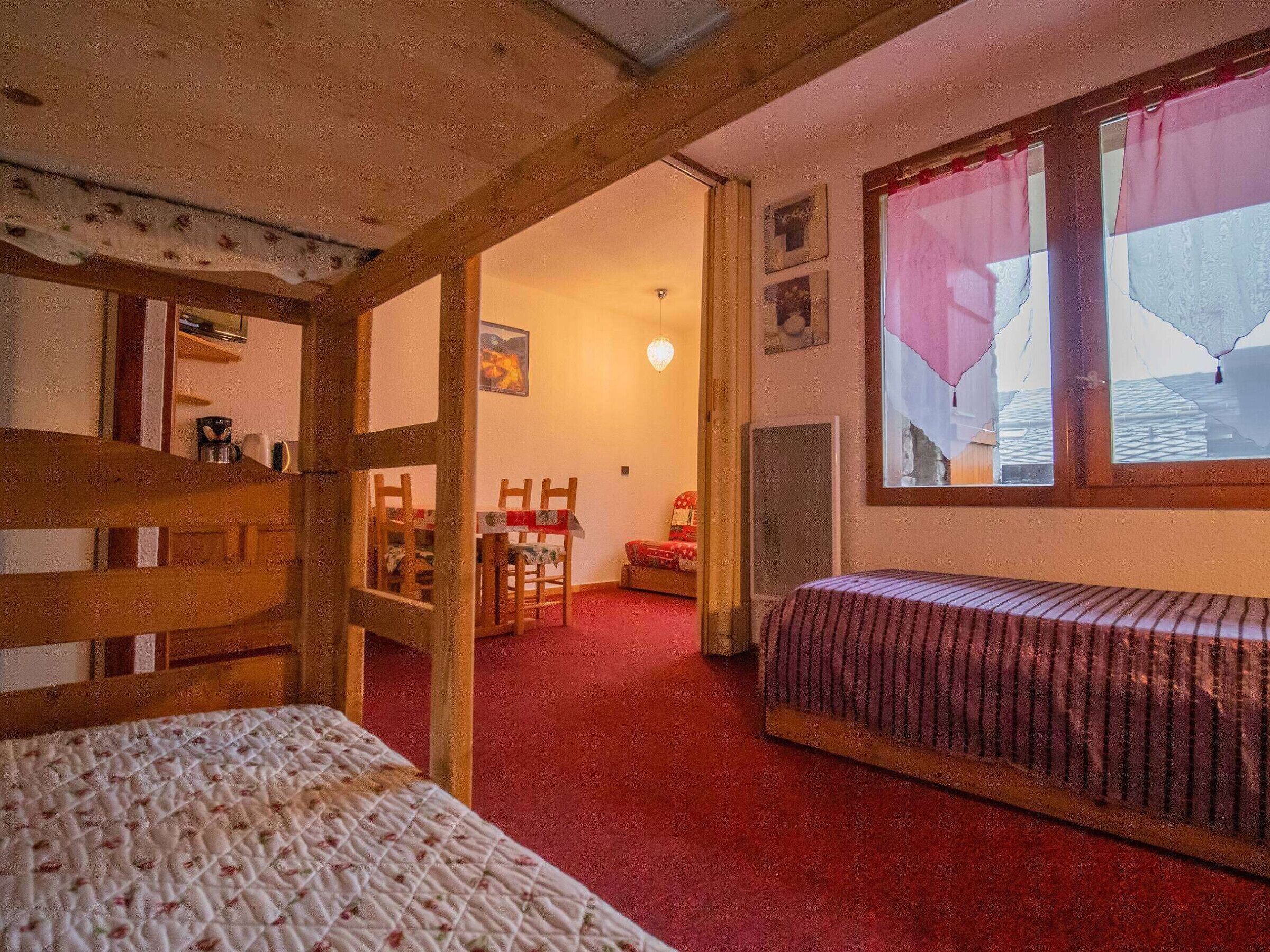 Ein gemütliches Zimmer mit einem Holzbettgestell, einem Einzelbett, einem roten Teppich, einem Essbereich mit Stühlen und einem großen Fenster mit Vorhängen.
