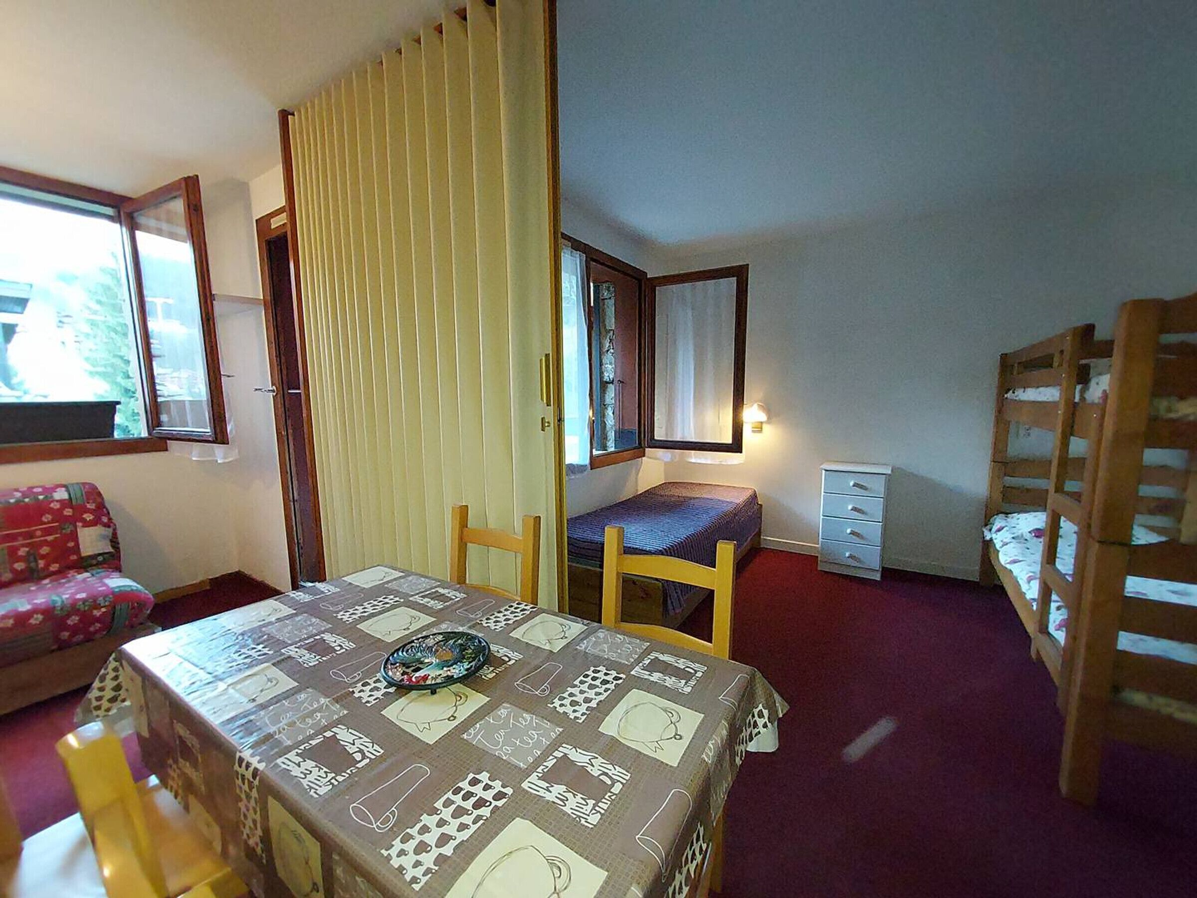 Et lille værelse med en seng, et skrivebord, en stol og et bord med et mønstret duge.