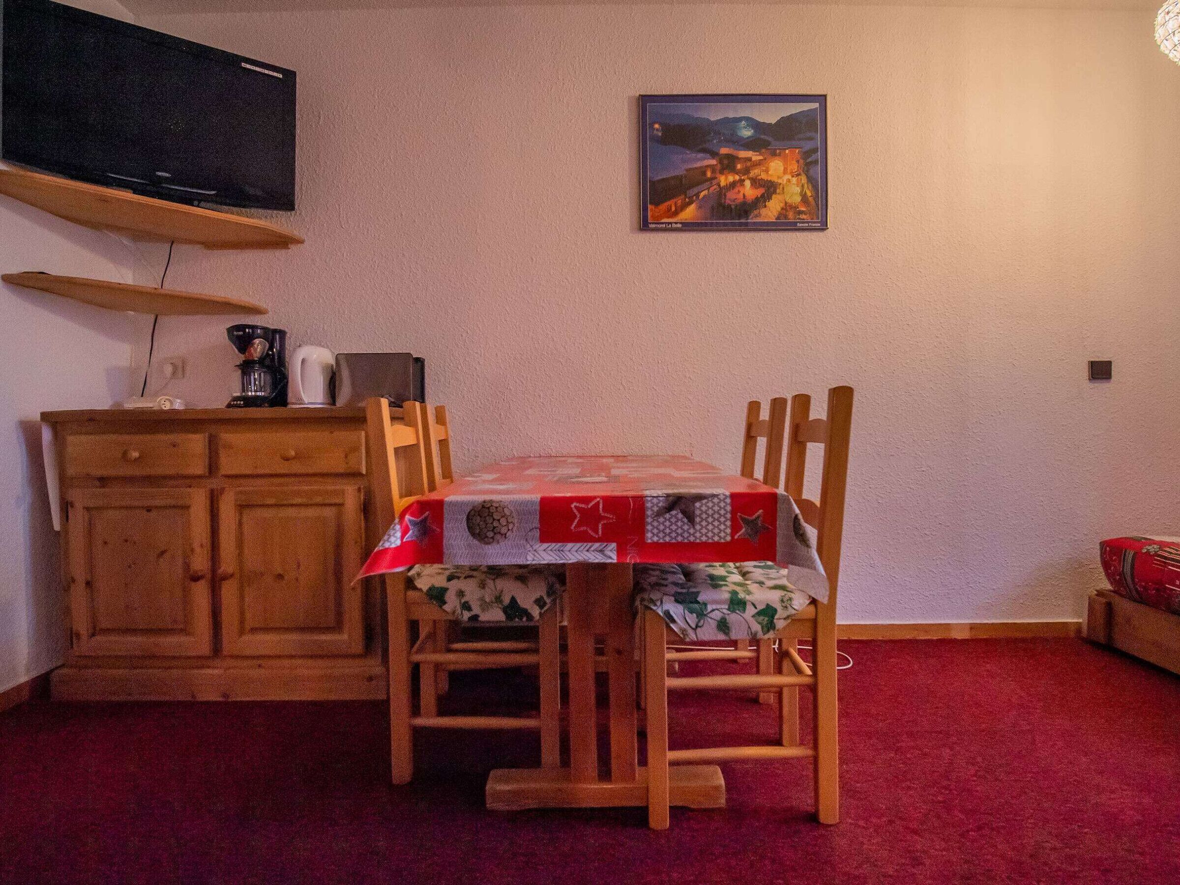 Ein Essbereich mit Tisch, bedeckt mit einer roten Tischdecke, Holzstühle, ein Holzschrank, ein Fernseher auf einem Schrank und ein gerahmtes Bild an der Wand.