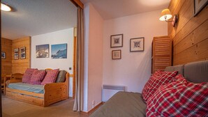 Ein gemütliches Zimmer mit einem hölzernen Bett, einer Holzbank und einem kleinen Tisch. An den Wänden hängen Bilderrahmen und eine Lampe ist angebracht.