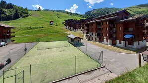 Blick auf ein Hotel mit mehreren Holzbauten, einem Tennisplatz und einer hügeligen Landschaft im Hintergrund.