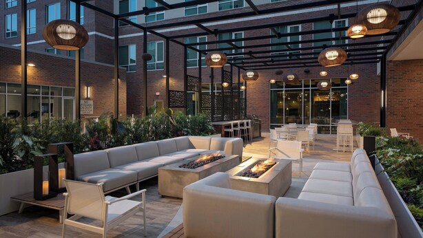 An outdoor patio with a modern design, featuring a seating area with sofas and a fire pit, surrounded by plants and under a pergola with hanging lights. - Residence Inn by Marriott Greenville Downtown (Greenville)