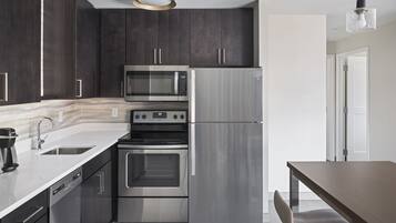 A modern kitchen with dark cabinets, stainless steel appliances, a dishwasher, and a dining area with a wooden table and chair.