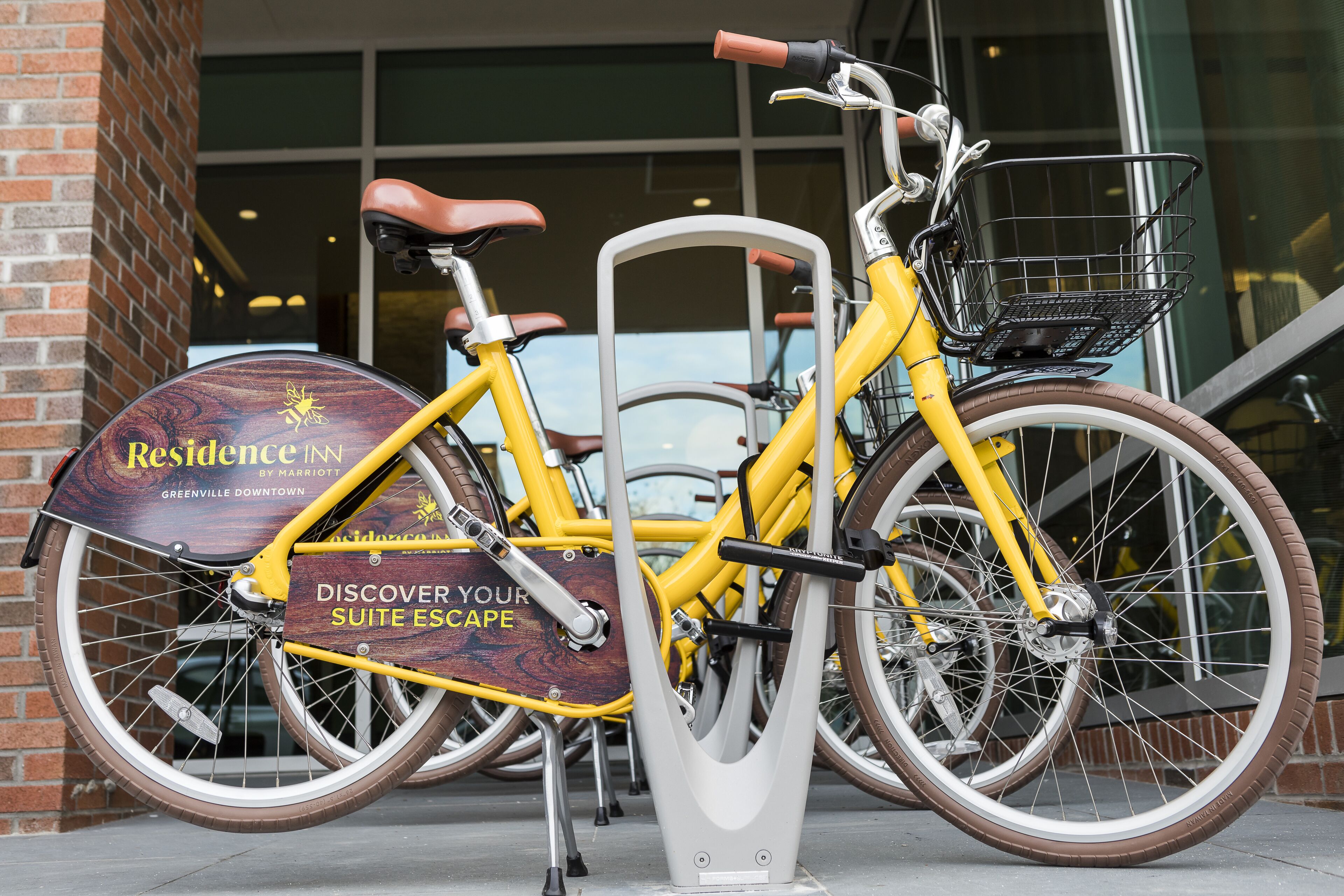 A yellow bicycle with a brown seat and a metal basket, parked in front of a building with a sign that reads "Resilience Inn by Marriott Greenville Downtown."