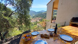 Un tavolo da pranzo in legno apparecchiato con piatti blu, posate, una bottiglia di vino e un vasetto di marmellata, con vista panoramica su un villaggio e montagne.