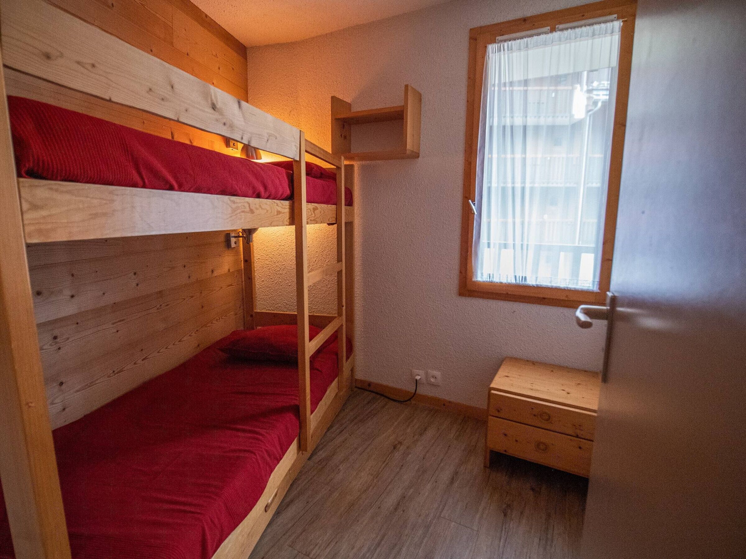 Dormitori amb lliteres, sòl de fusta, una petita taula de nit de fusta i una finestra amb cortines.