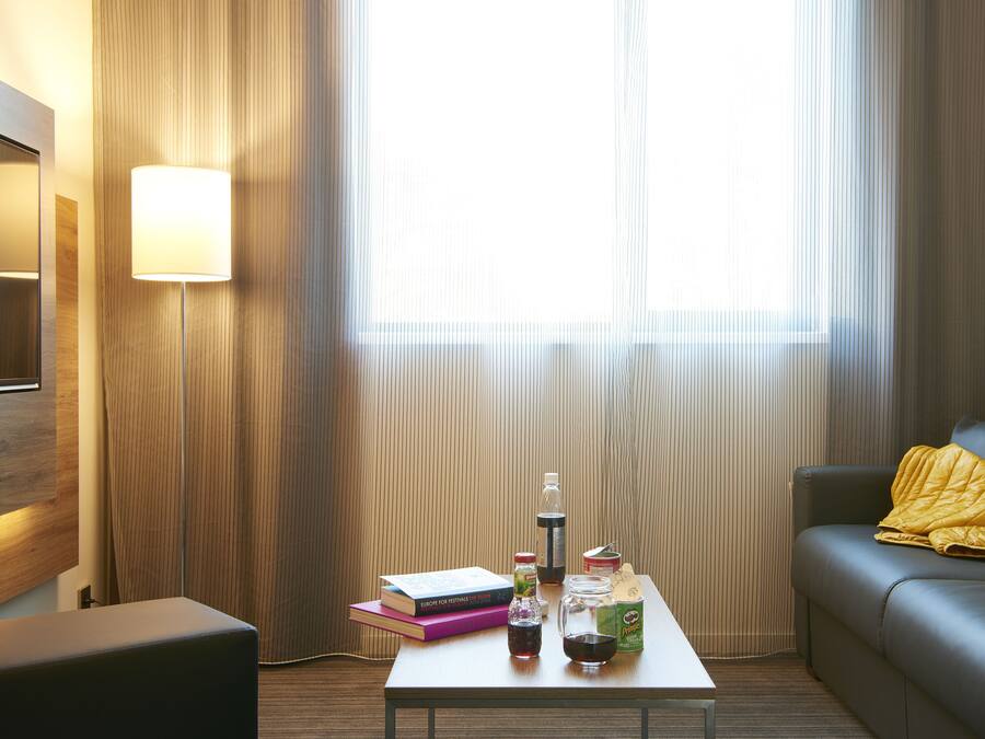 A hotel room with a sofa, a coffee table with books, a lamp, and a window with curtains.