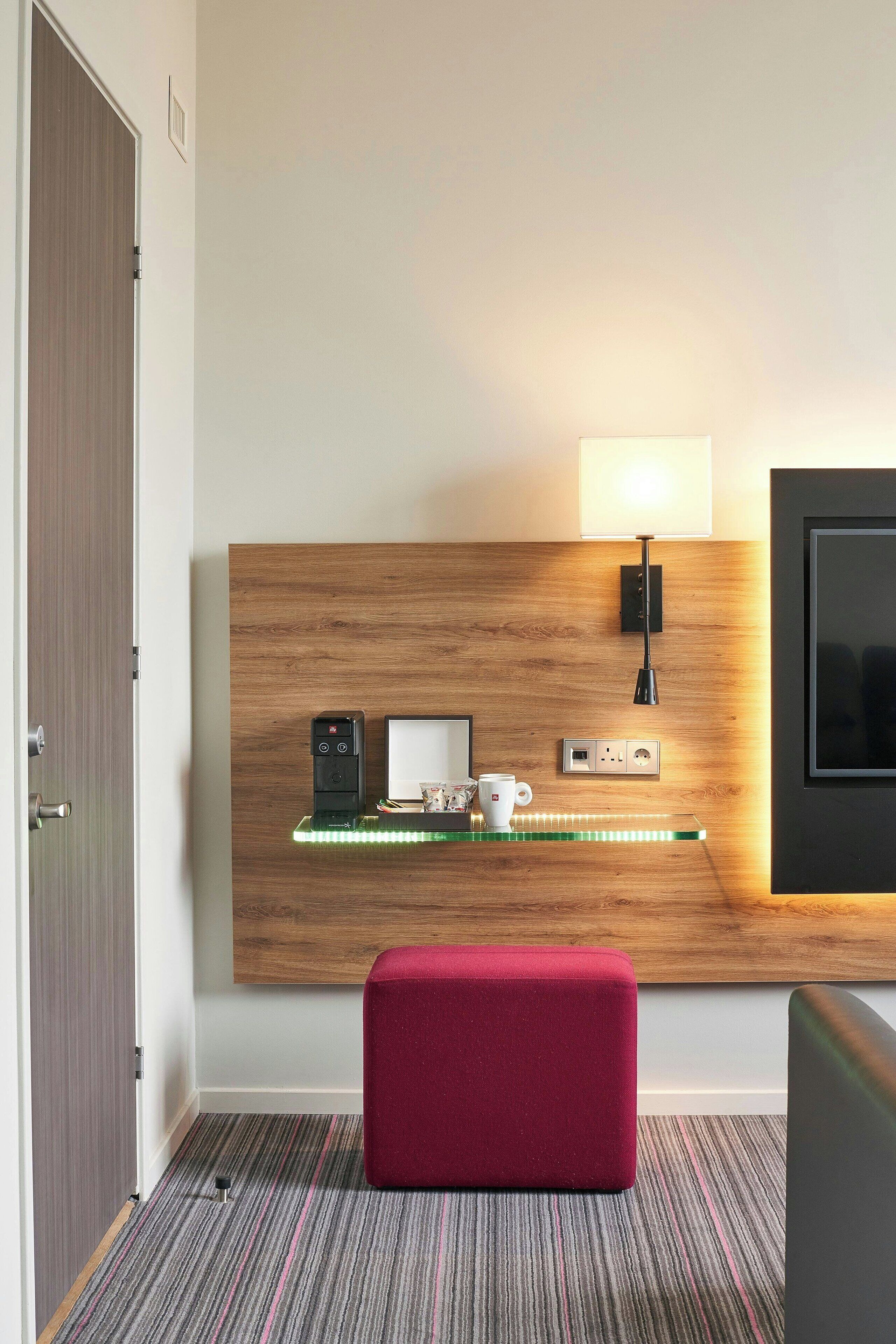 Habitación de hotel con cabecera de madera, mesita de noche con lámpara, reposapiés rojo y televisor en la pared.