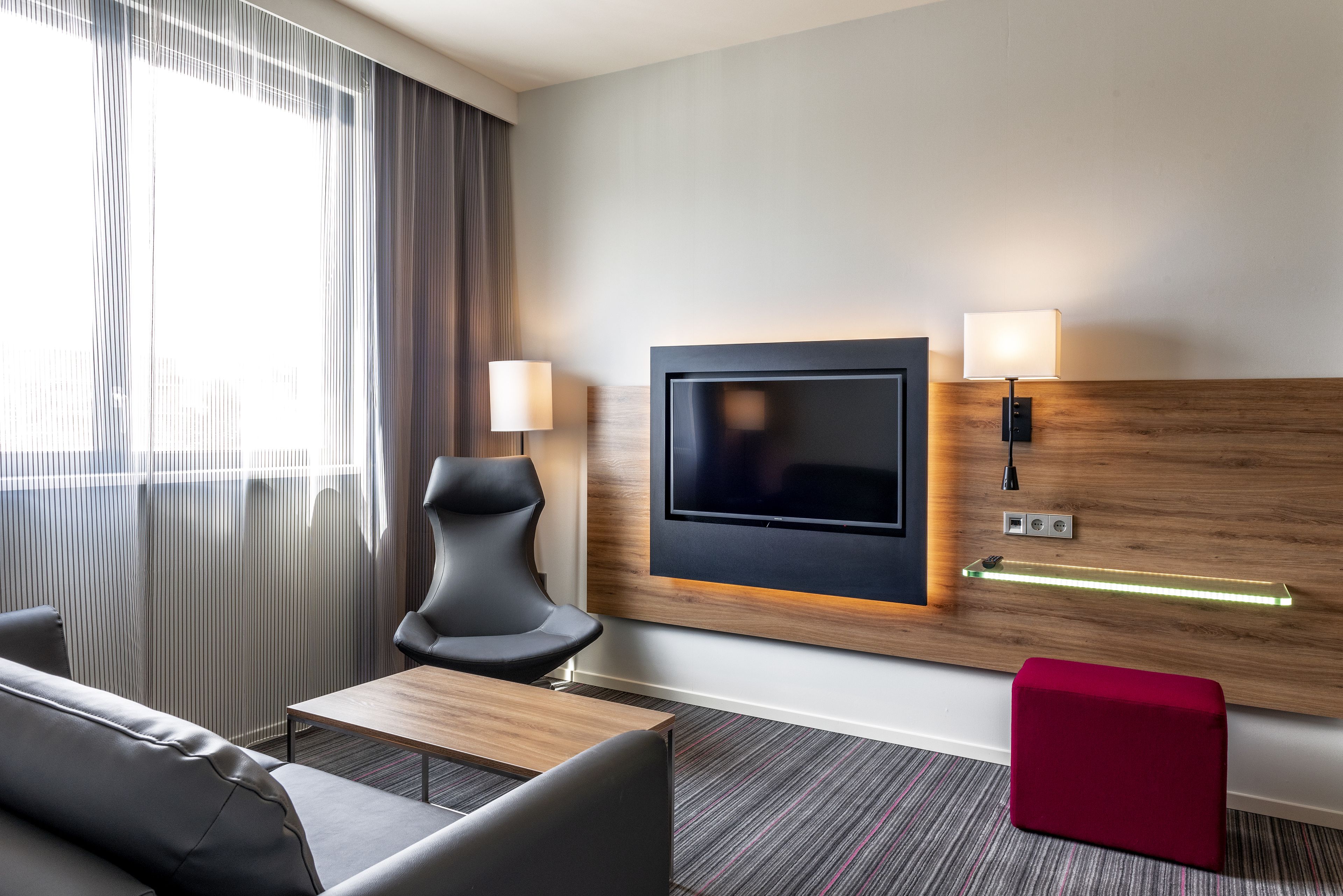 A hotel room with a TV mounted on the wall, a sofa, a chair, a small table, and a red ottoman.
