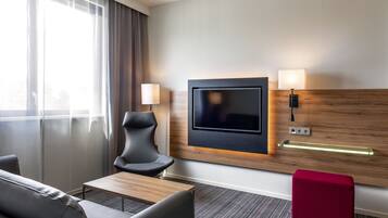 A hotel room with a TV mounted on the wall, a sofa, a chair, a small table, and a red ottoman.