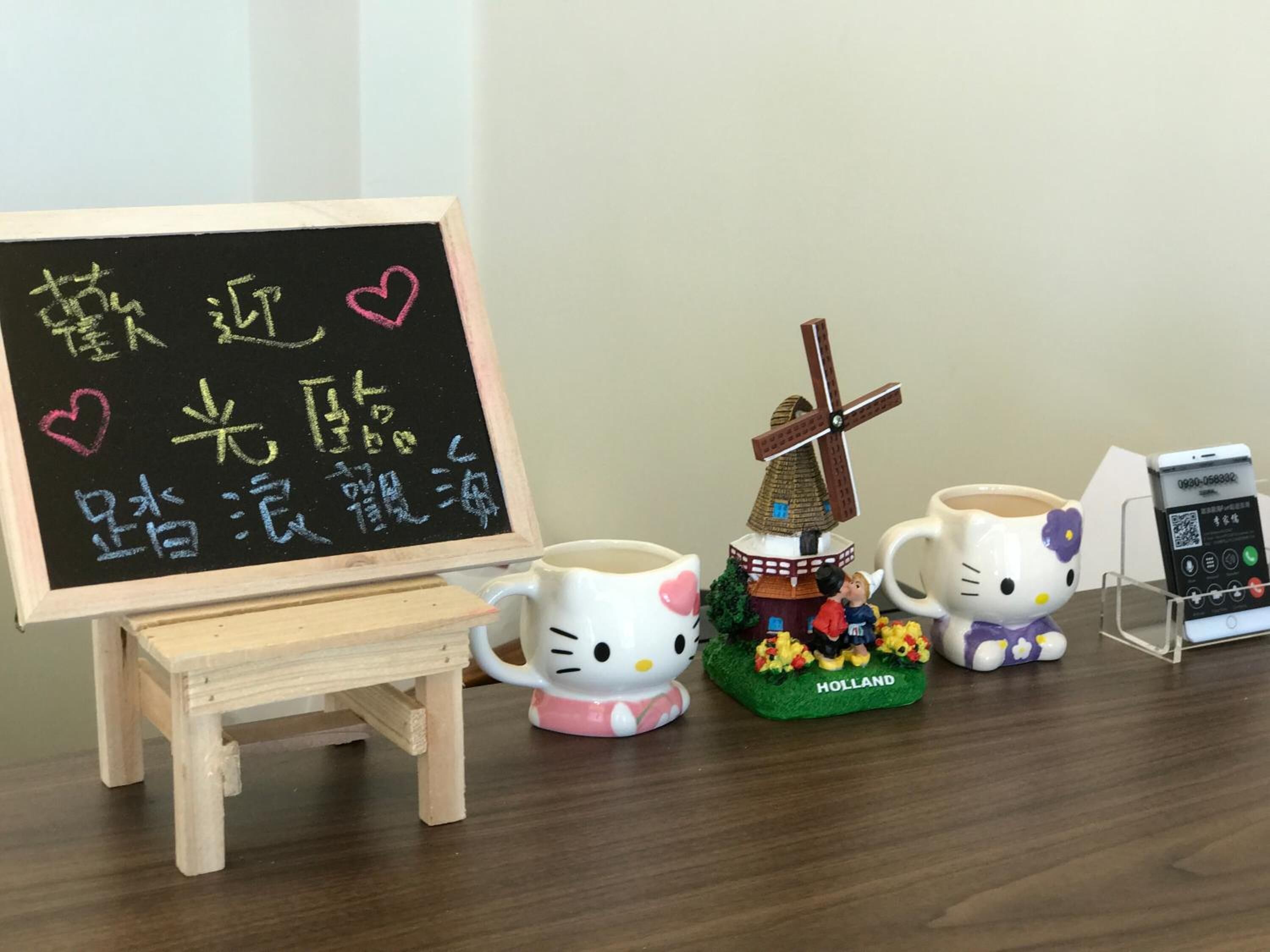 Un petit présentoir en bois sur lequel sont posés un tableau noir avec des inscriptions et des cœurs, deux tasses à l’effigie de Hello Kitty, une figurine de moulin à vent et un lecteur de codes QR.