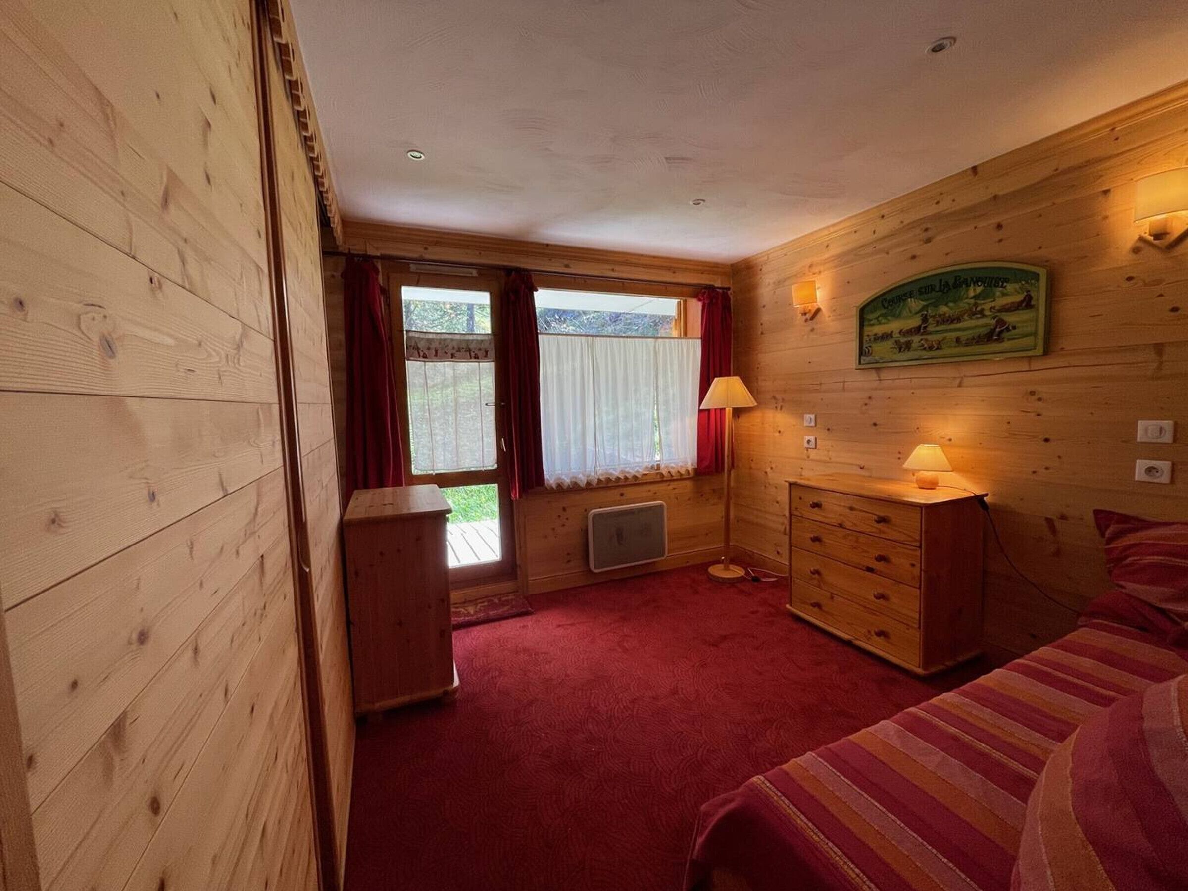 A wooden room with a bed, a dresser, a radiator, and a window with curtains.