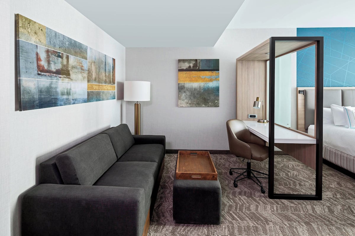Una habitación de hotel moderna con un sofá gris, una mesita de madera, un escritorio con una silla, un espejo grande y obras de arte abstractas en las paredes.