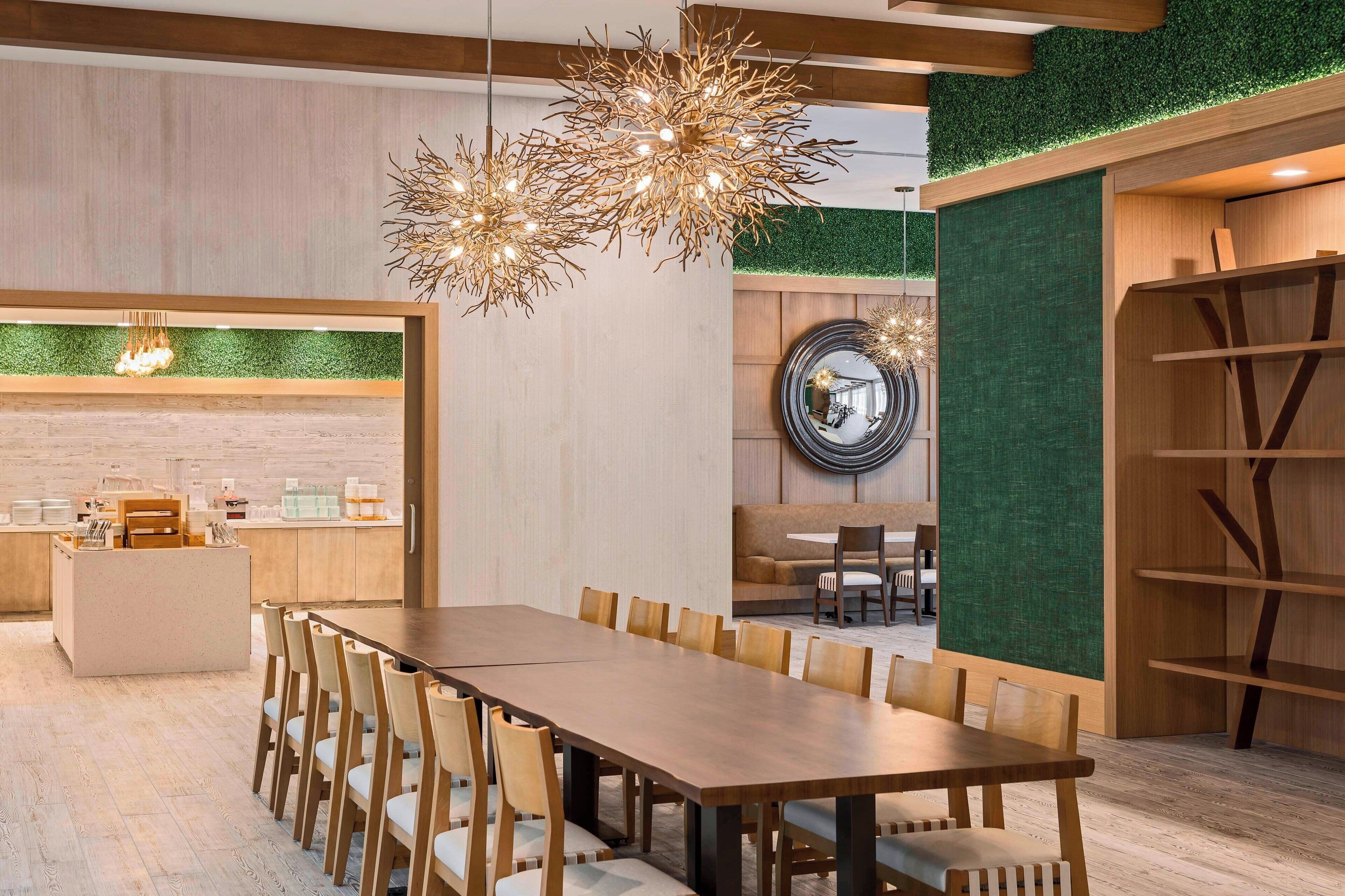 A modern dining area with a long wooden table and chairs, a unique chandelier, and a green wall with a mirror.