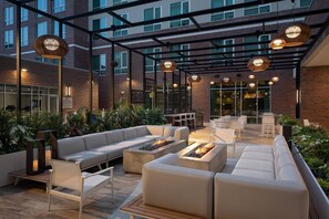An outdoor patio with a modern design, featuring a seating area with sofas and a fire pit, surrounded by plants and under a pergola with hanging lights.