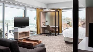 A hotel room with a large window, a desk, a chair, a bed, and a TV.