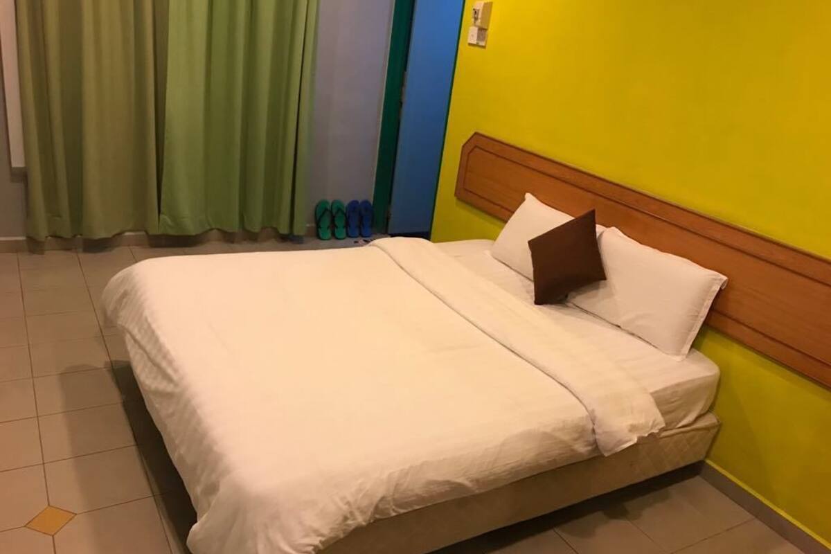 Una cama con ropa de cama blanca, un almohón marrón y un cabecero de madera en una habitación con paredes amarillas y cortinas verdes.