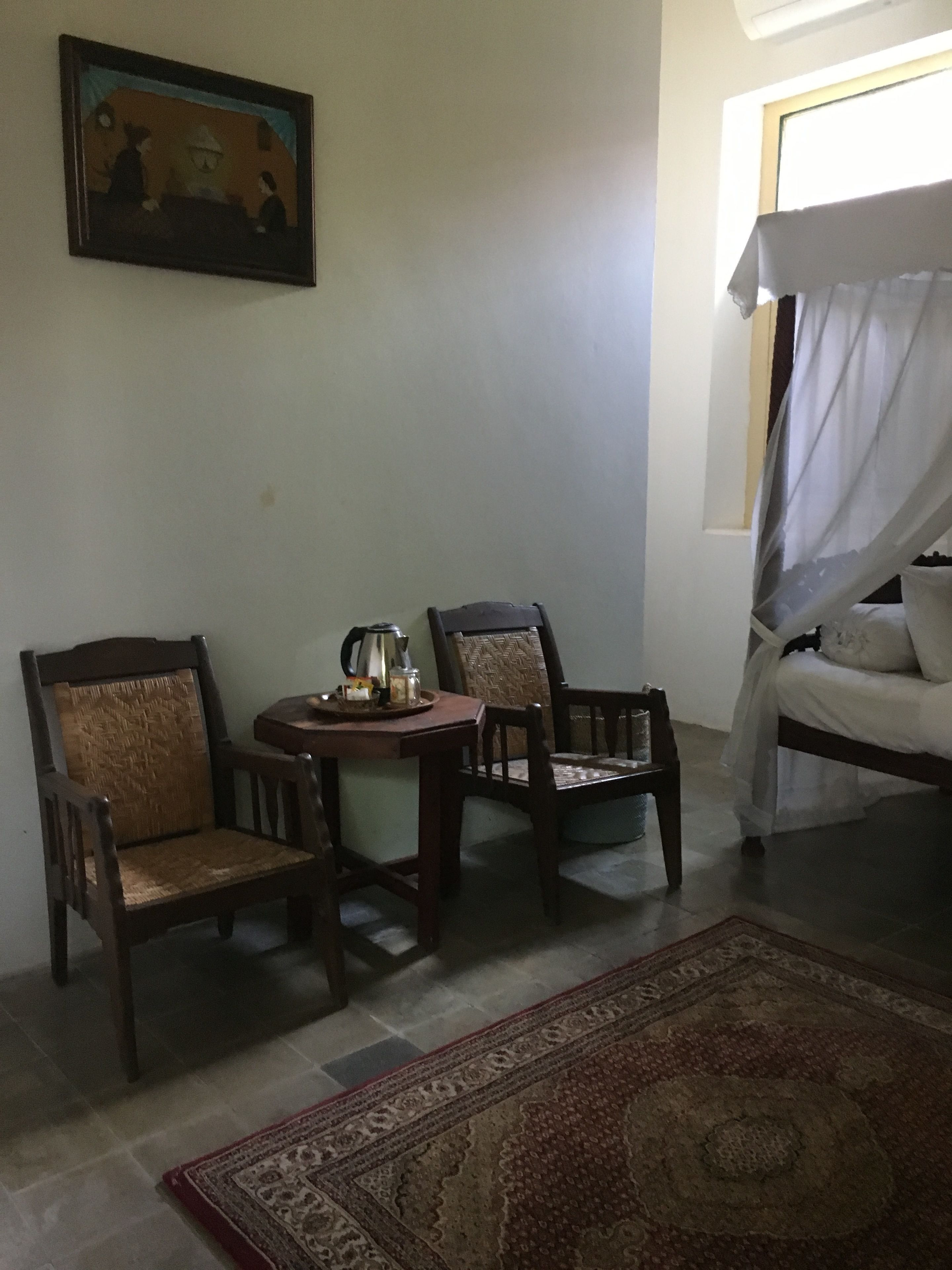 Una habitación con dos sillas de madera, una mesita redonda con una tetera, una cama con dosel y una alfombra estampada en el suelo.