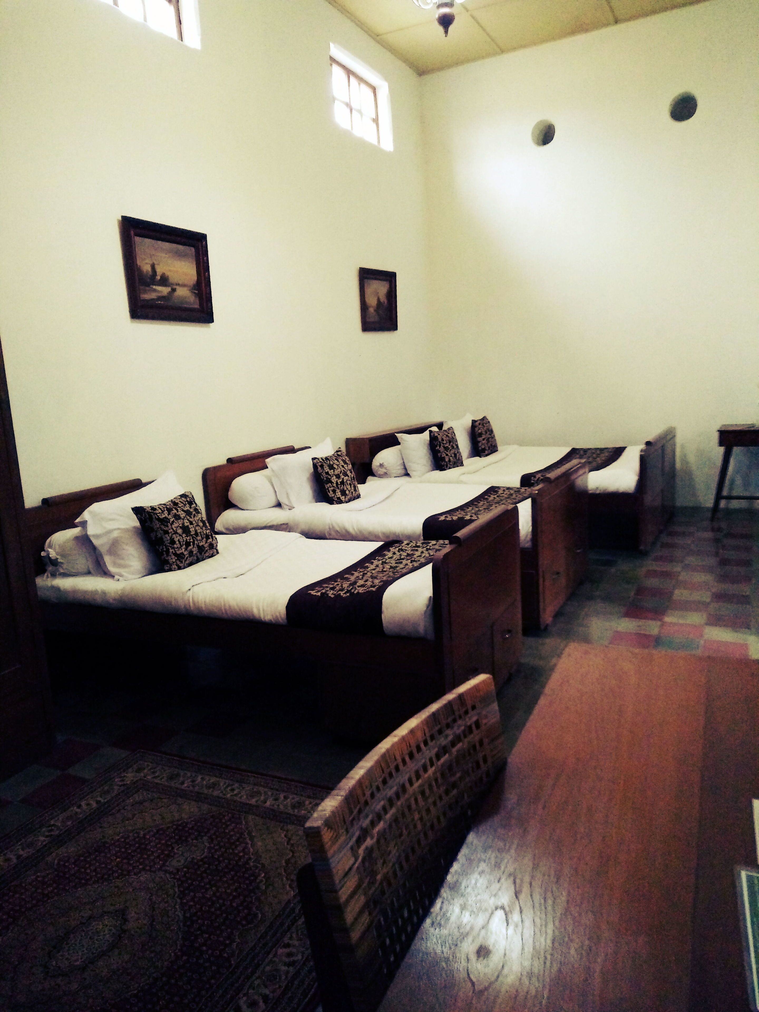 A room with four single beds aligned in a row, each with a wooden headboard and white bedding. There are two windows on the wall, one with a view of the outside. A patterned rug is on the floor, and a wooden bench is in the foreground.