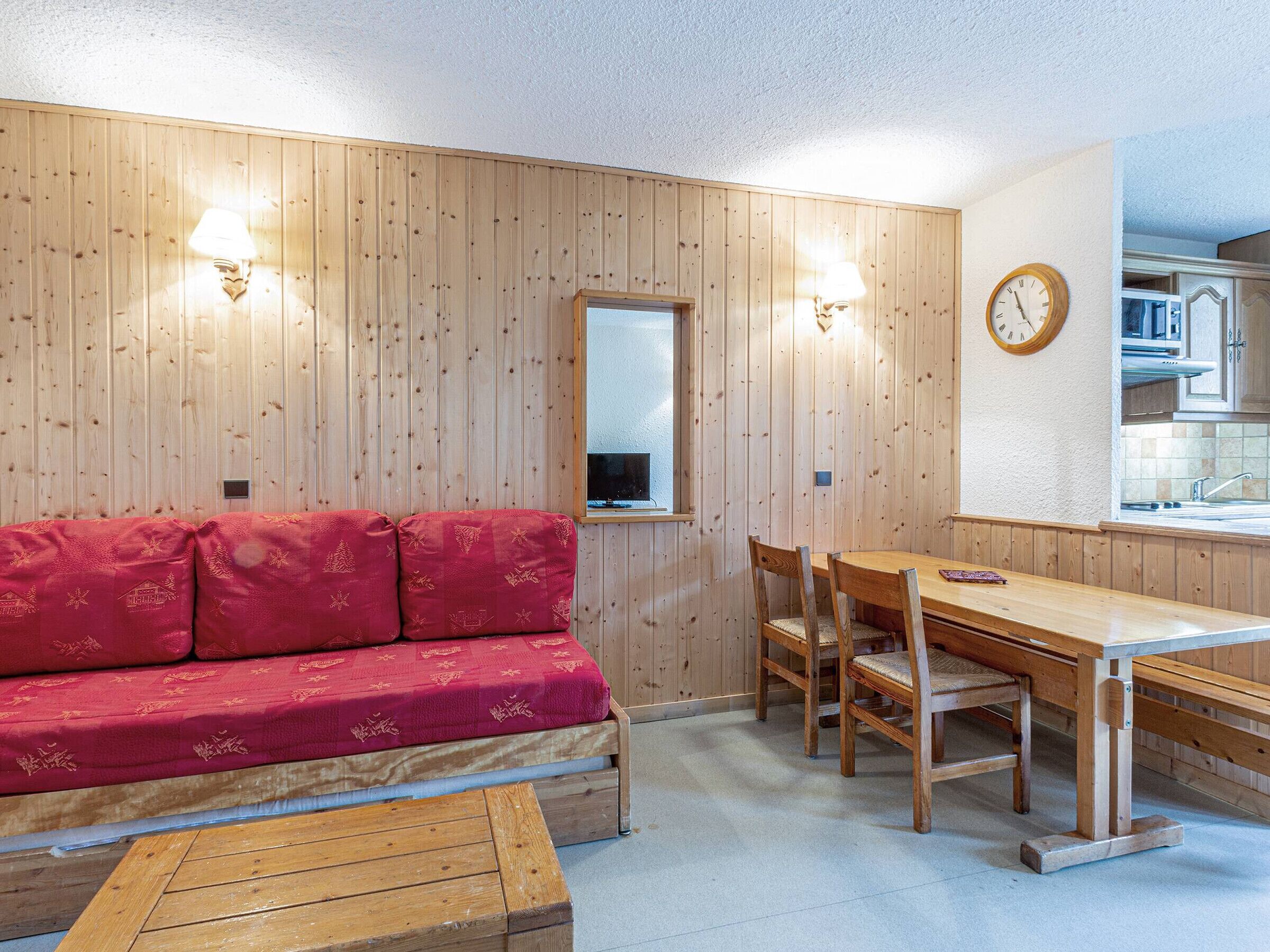 Una habitación acogedora con pared de madera, un sofá rojo, una mesa de comedor de madera con sillas y una cocina visible a través de una puerta abierta.