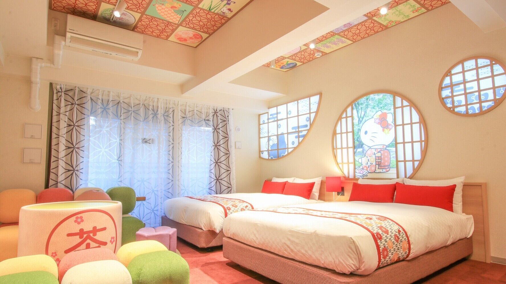 A hotel room with two beds, a large window with a Hello Kitty design, a ceiling with a geometric pattern, and a small seating area with colorful cushions.