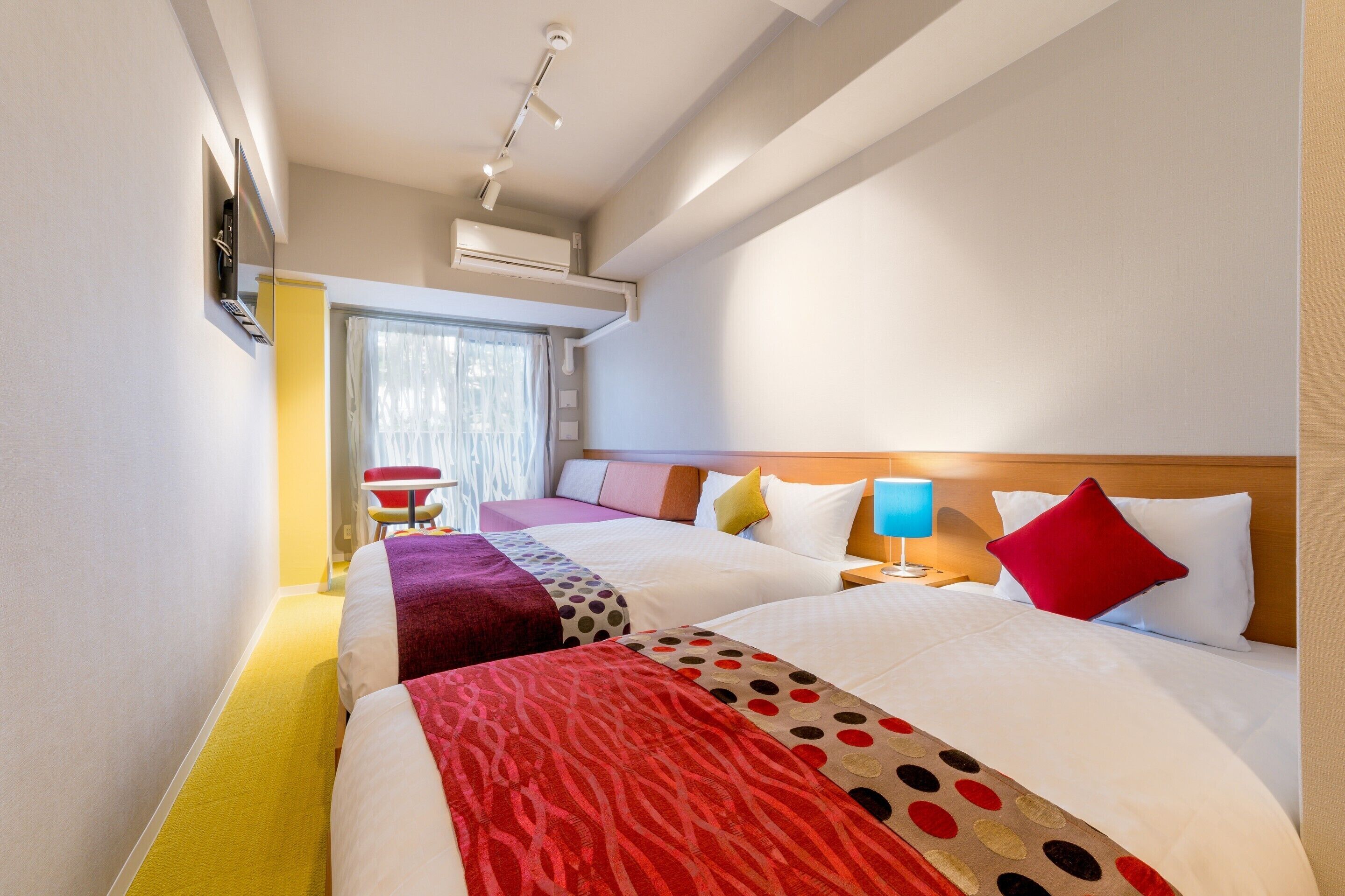 A hotel room with two beds, a window with curtains, an air conditioning unit, a red chair, and a nightstand with a lamp.