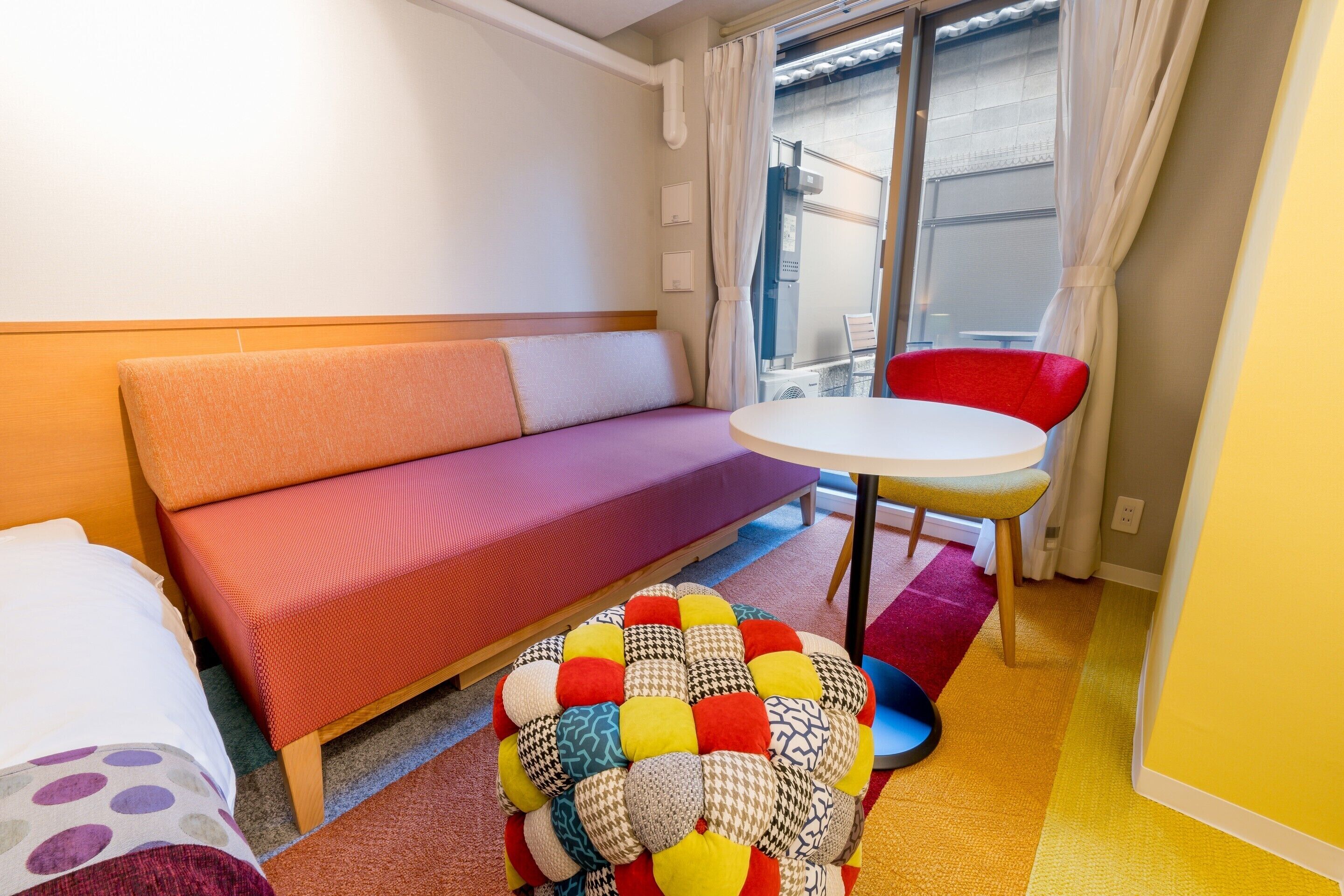Una habitación con sofá, una mesita redonda, un cojín de colores y una ventana con cortinas.