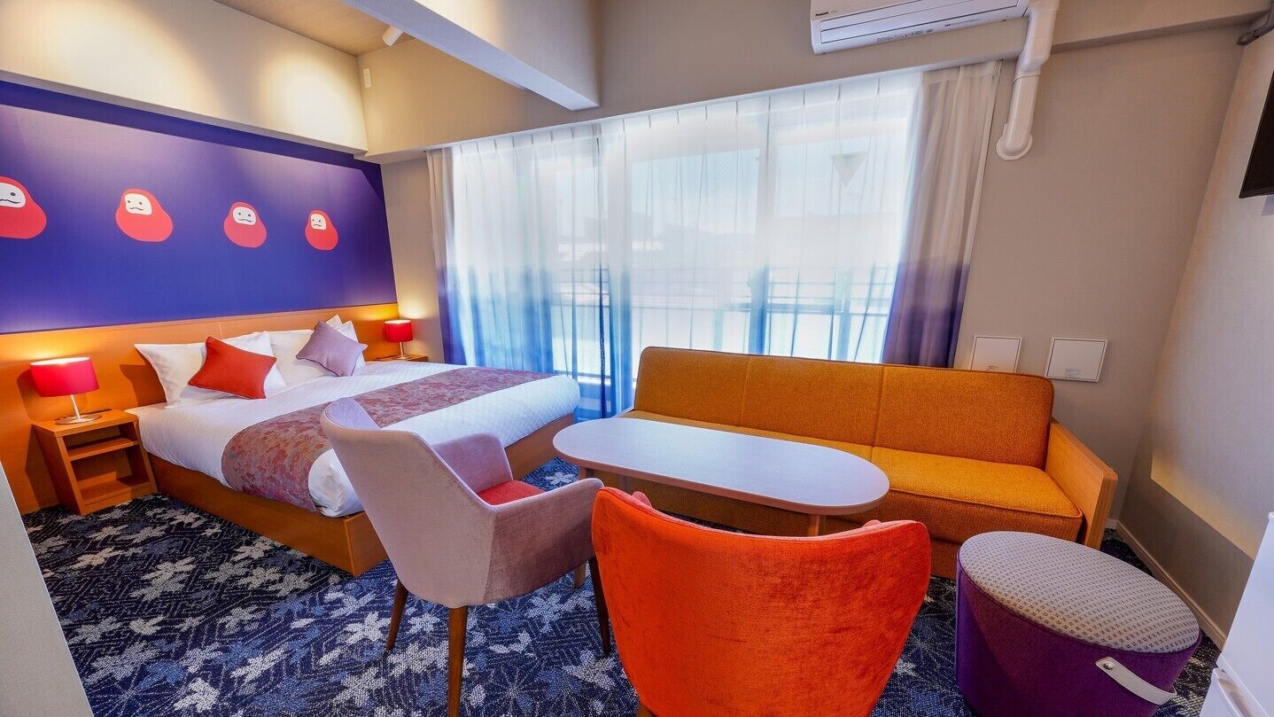A hotel room with a bed, a sofa, a small table, a chair, and a window with curtains.