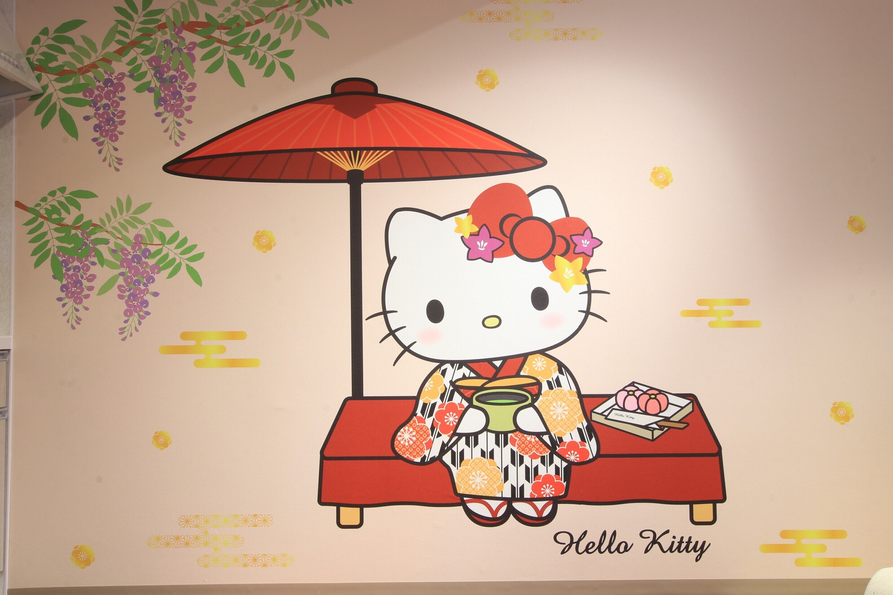 A wall with a Hello Kitty illustration, a red umbrella, and a table with a teapot and a plate of food.