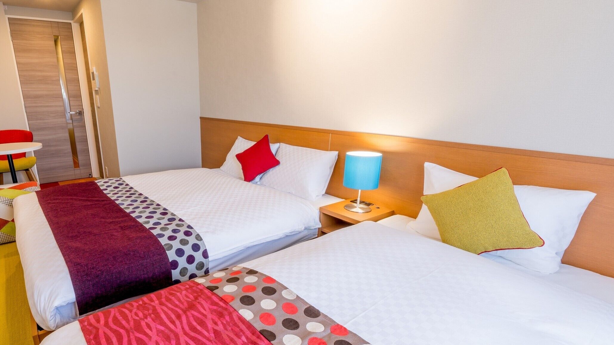 A hotel room with two beds, a nightstand with a lamp, a table with chairs, and a door.