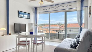 A balcony with a view of buildings, a ceiling fan, a TV, and a sofa with a striped pillow. - Cozy Appart 150 m from the beach with balcony + Parking (Biscarrosse)