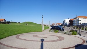 A paved area with a circular design, a road with parked cars, and buildings with red roofs in the background. - Cozy Appart 150 m from the beach with balcony + Parking (Biscarrosse)