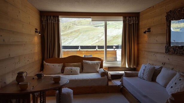 A cozy room with a bed, a sofa, a table with a mug, and a view of a hillside through large windows.
