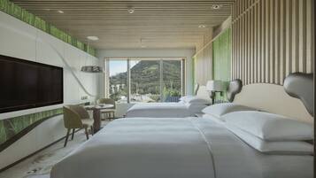 A modern hotel room with a large bed, a TV, a desk, and a view of a mountainous landscape.