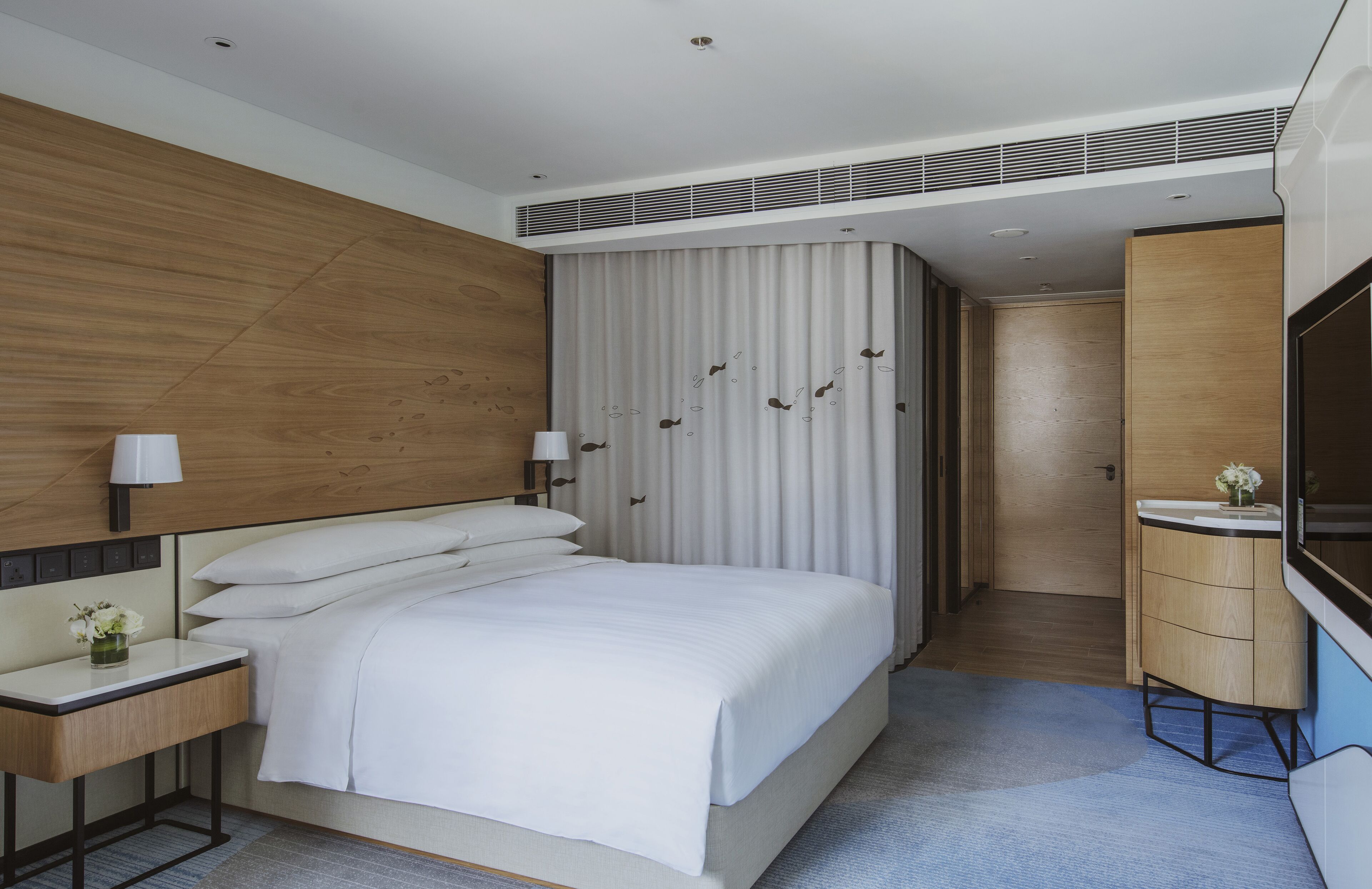 Een moderne hotelkamer met een groot bed, nachtkastjes, een televisie, een houten kledingkast en een badkamer met een wastafel en spiegel.